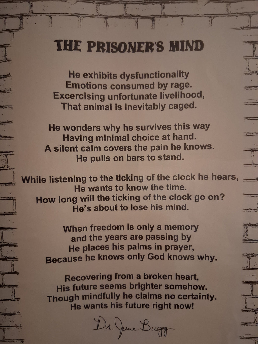 A printed poster or sign with the title 'THE PRISONER'S MIND' and a long poem in black text on an off-white background, mounted against a faux brick wall. Signed with the alias name, ''Dr. June Bugg.'' The author is Eddie Wyatt Jr.