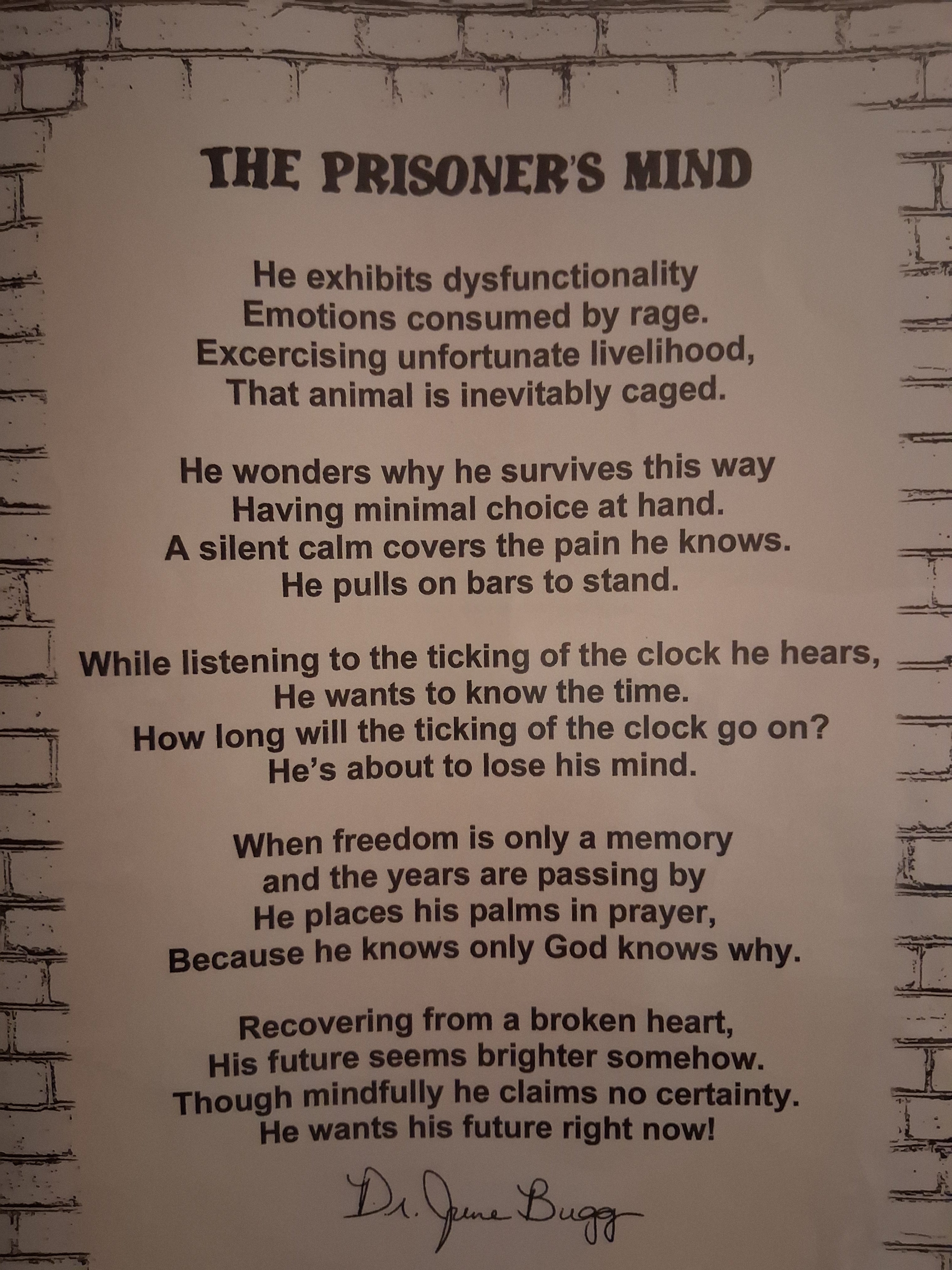 A printed poster or sign with the title 'THE PRISONER'S MIND' and a long poem in black text on an off-white background, mounted against a faux brick wall. Signed with the alias name, ''Dr. June Bugg.'' The author is Eddie Wyatt Jr.
